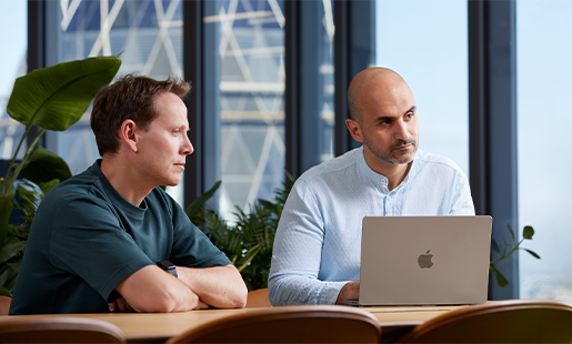 أحمد، أمامه جهاز MacBook، ويجلس أمام طاولة مع زميل له وينظر إلى شخص خارج نطاق الصورة.
