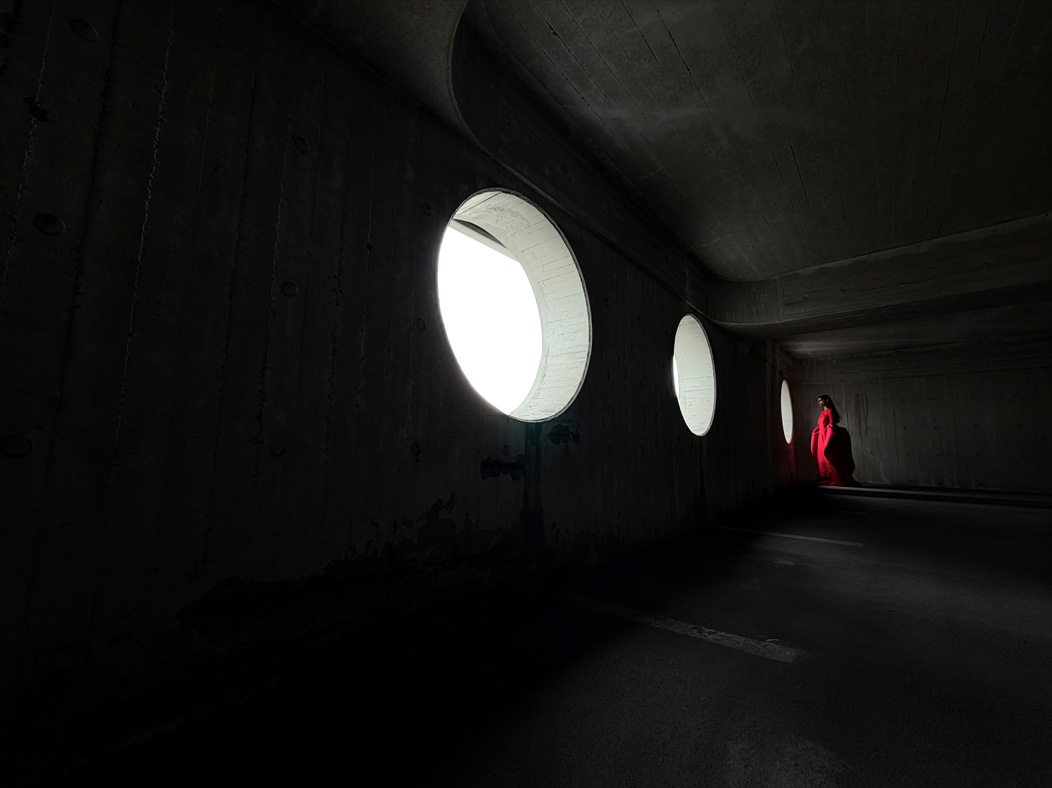 Žena v dlouhých červených šatech stojí při fotografování v prázdné místnosti. Aplikace Fotky přepíná mezi ohniskovými vzdálenostmi od 200 mm do 13 mm na ukázku možností profesionální fotosoustavy.